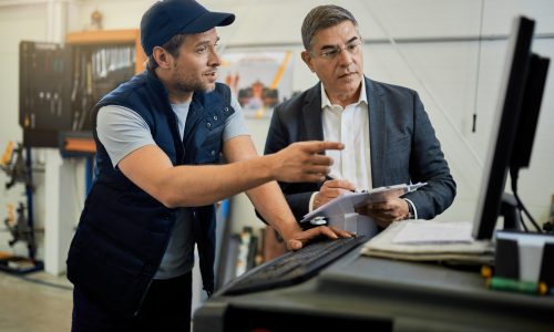 Mid adult manager and car mechanic talking while using computer in auto repair shop.